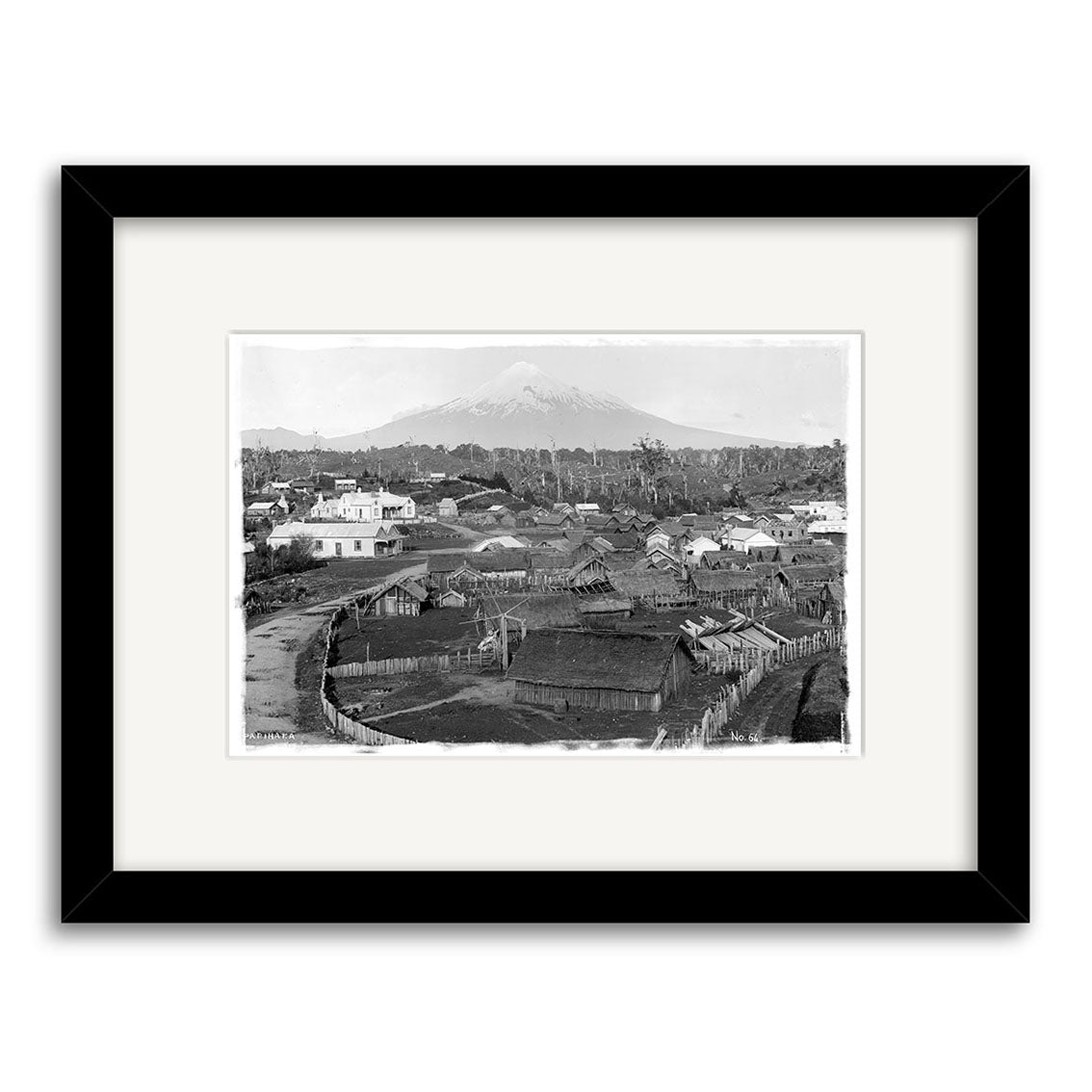 Mount Taranaki 1890 - HI142 is a black and white historical photographic print of a small town with wooden fences and buildings, set against a snow-capped mountain. Printed on matte paper, it comes framed with a white mat and black frame.
