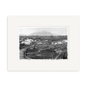 A black-and-white historical photographic print, Mount Taranaki 1890 - HI142, shows a village with thatched and wooden buildings in the foreground and a snow-capped mountain behind, framed by archival quality matte paper.