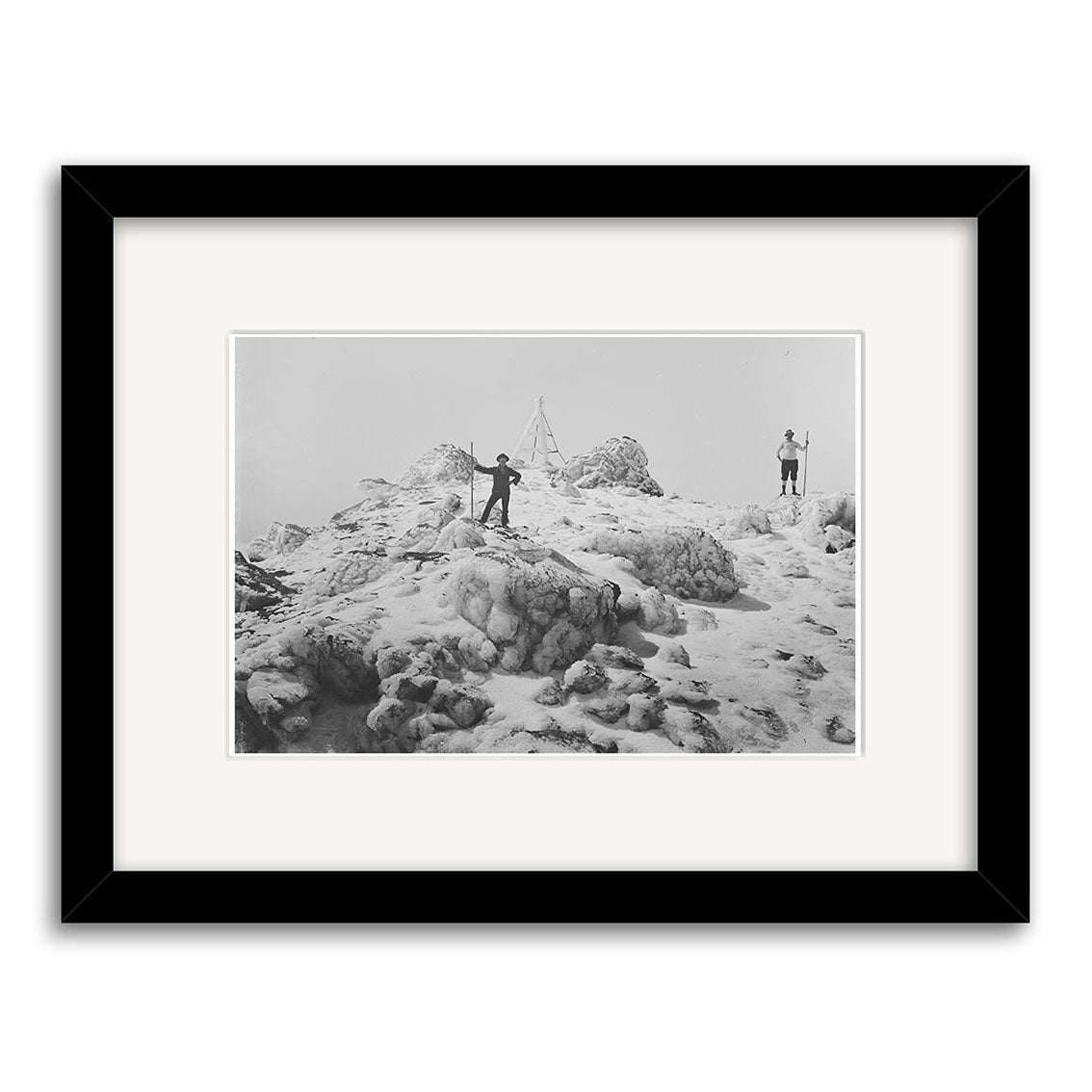 Mt Egmont 1911 - HI143: Black and white photo of two people on a snowy New Zealand mountain summit near a metal triangular structure; one raises an arm, the other with a walking stick. Framed with archival quality matte paper.