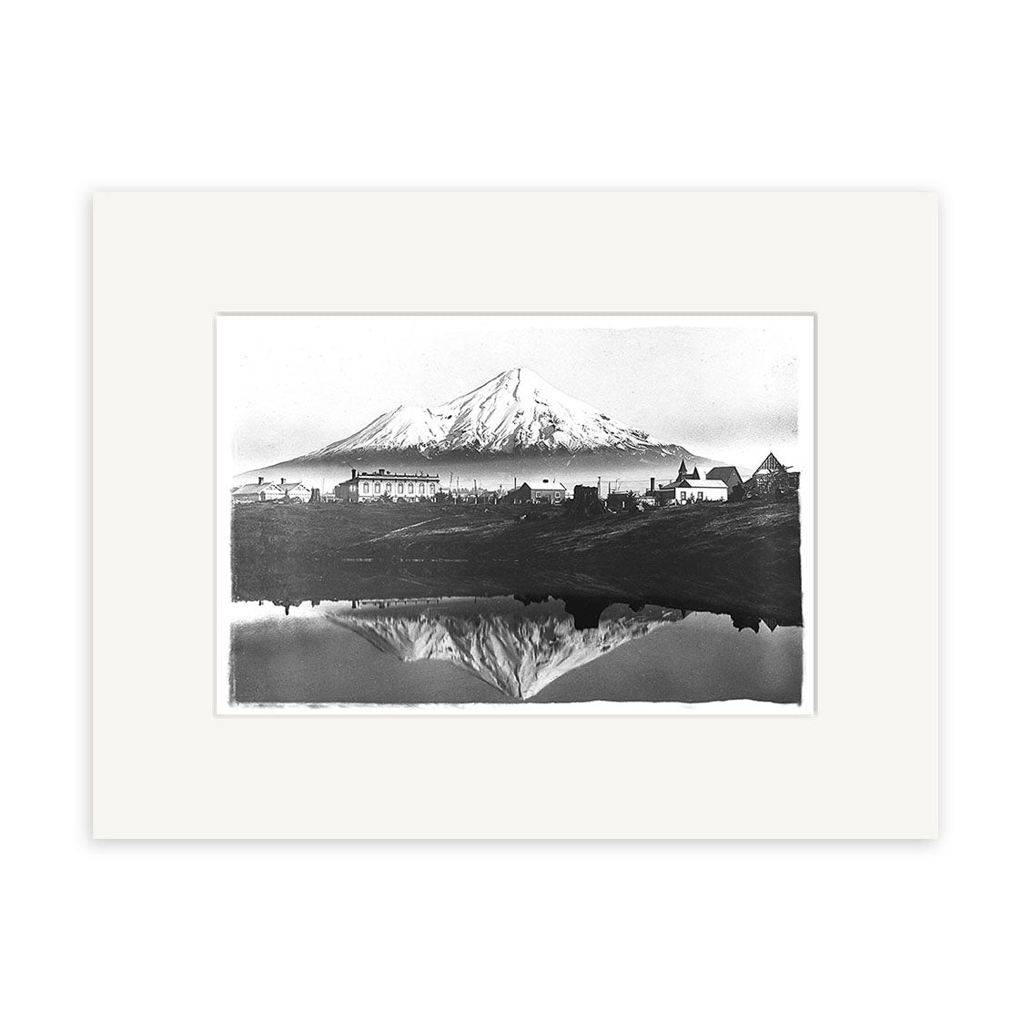 The Mt Taranaki 1900 - HI144 is a black and white illustration of New Zealand’s snow-capped Mt Taranaki with buildings at its base, reflected in still water. This historical photographic print comes centered on an archival quality white mat.
