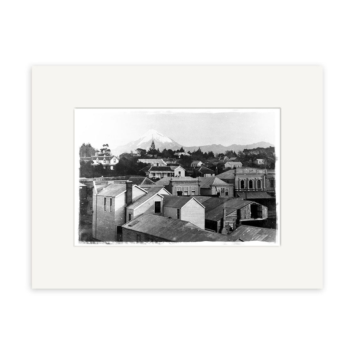 The Mt Taranaki 1881 - HI145 black and white historical photographic print features small town rooftops, wooden buildings, trees, and a mountain backdrop, framed in a white mat on archival quality matte paper.
