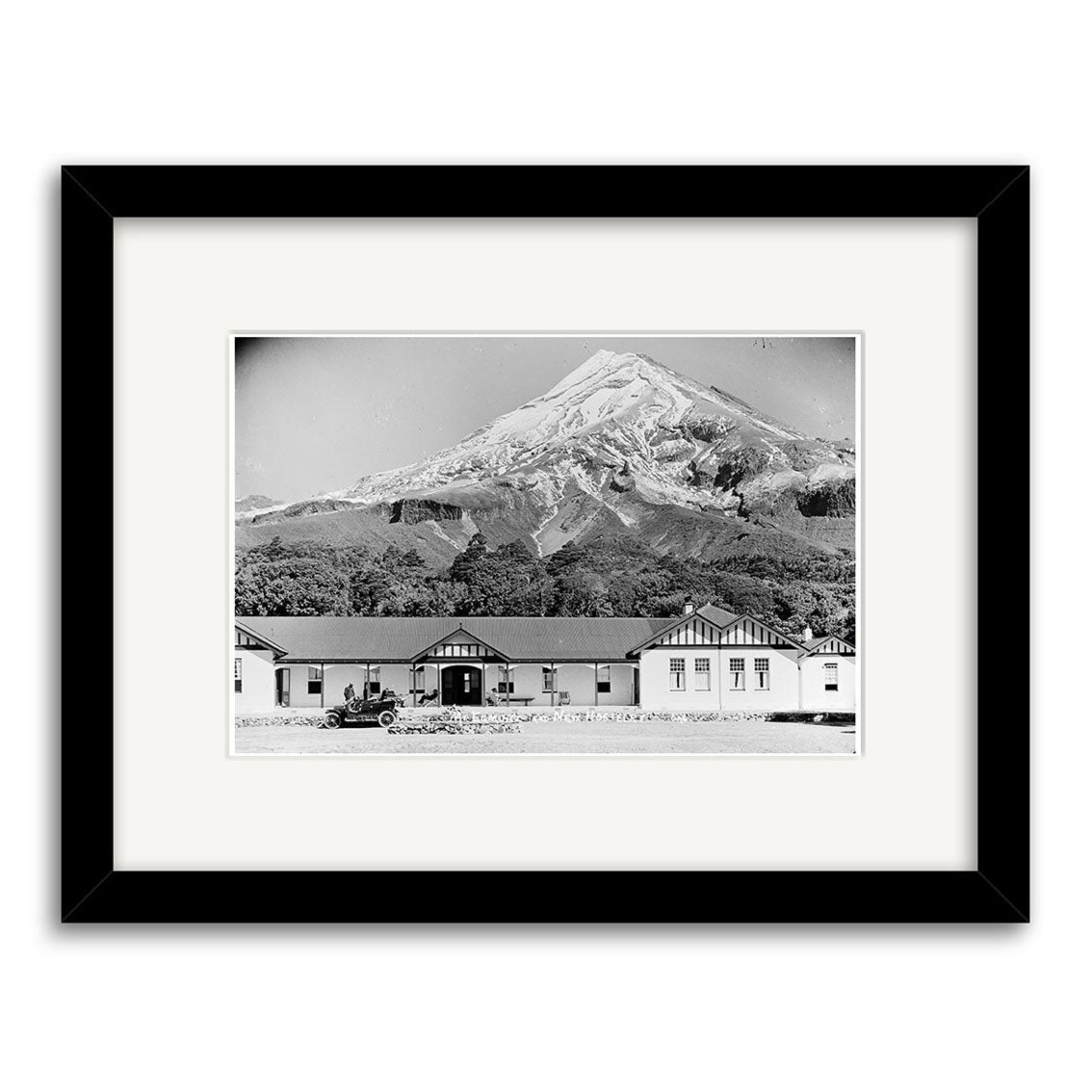 The North Egmont Hostel 1910 - HI147 features a black and white framed photo printed on archival matte paper, depicting single-story buildings, a vintage car, and a snow-capped mountain in the background.
