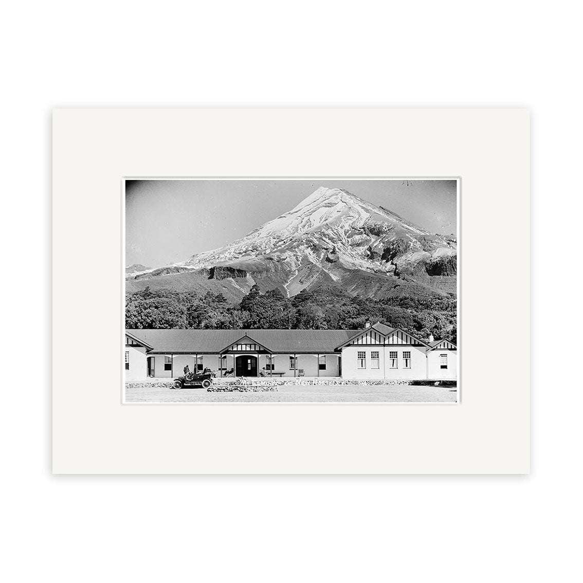 The North Egmont Hostel 1910 - HI147 print features a vintage car before three single-story buildings with snow-capped Mount Taranaki in the background. Printed in black and white with a white border on archival quality matte paper.