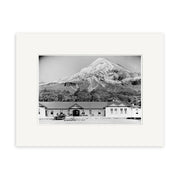 The North Egmont Hostel 1910 - HI147 print features a vintage car before three single-story buildings with snow-capped Mount Taranaki in the background. Printed in black and white with a white border on archival quality matte paper.