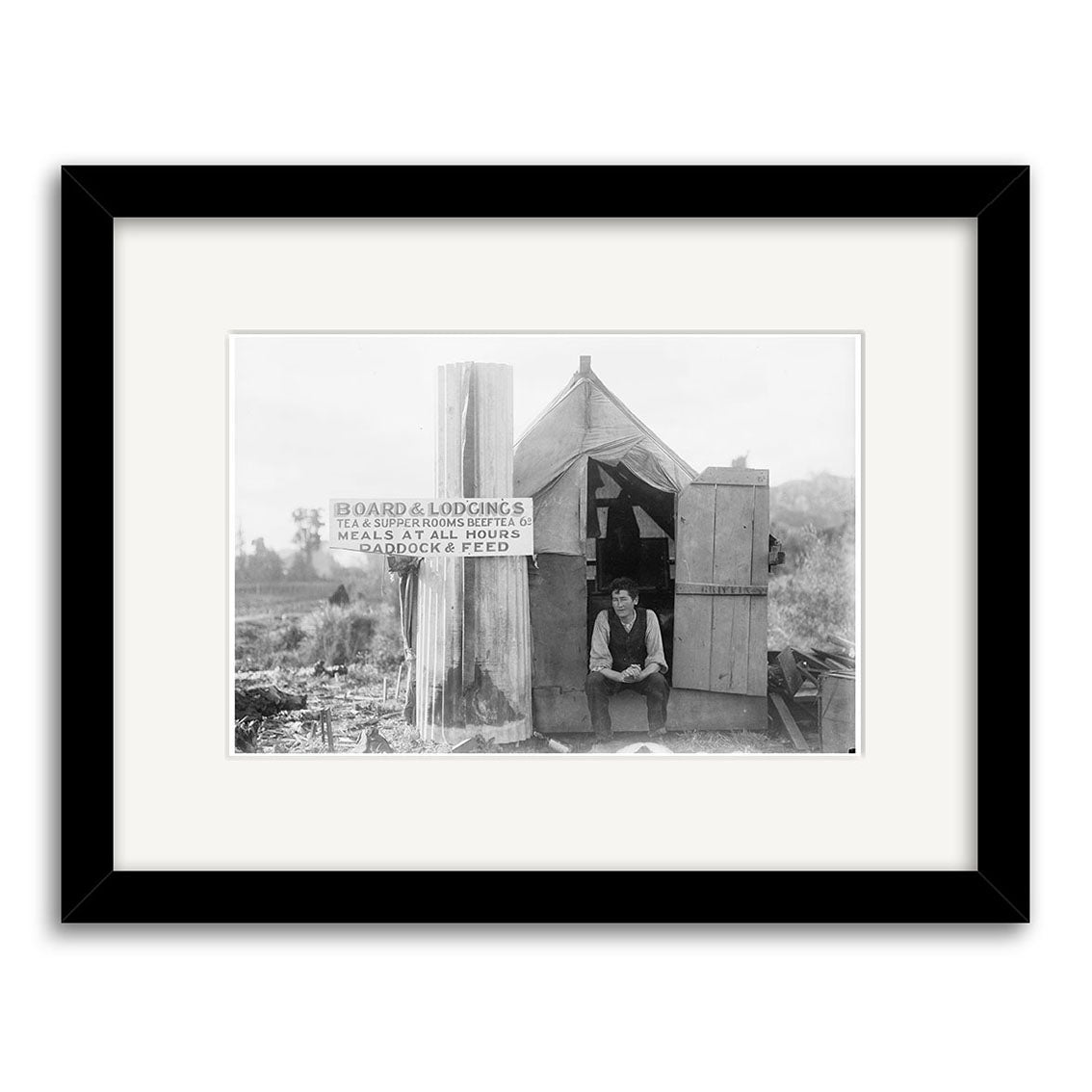 Small Hutt Taranaki 1910 - HI149: Black-and-white historical photographic print showing a man in front of a small wooden shack with a sign offering board, lodging, meals at all or regular hours, and paddock & feed. Printed on matte paper.