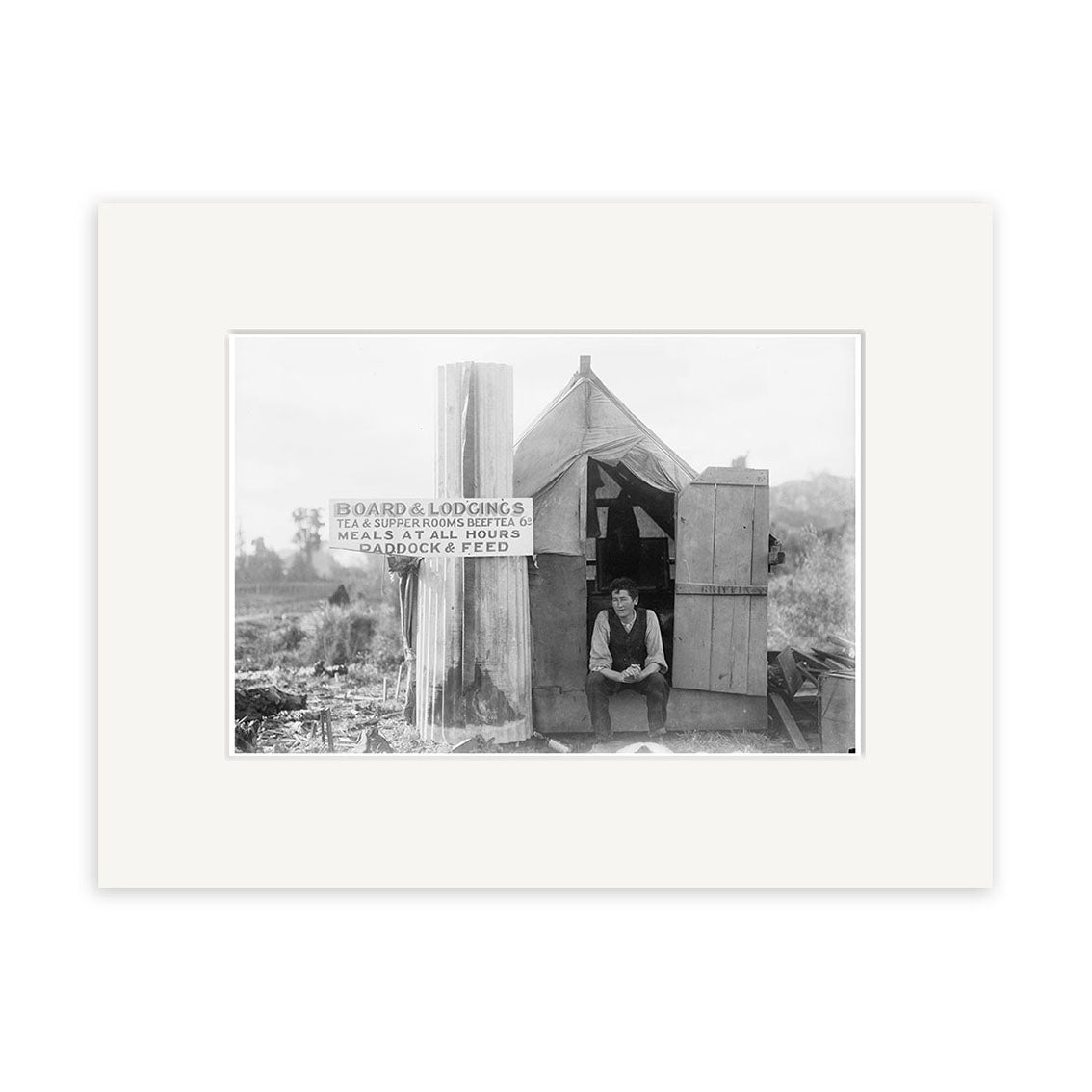 A man sits in the doorway of a small wooden shack beside a sign for board, lodging, meals, and paddock horses, captured in the Small Hutt Taranaki 1910 - HI149 Historical Photographic Print on matte archival paper.