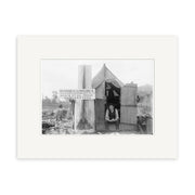 A man sits in the doorway of a small wooden shack beside a sign for board, lodging, meals, and paddock horses, captured in the Small Hutt Taranaki 1910 - HI149 Historical Photographic Print on matte archival paper.
