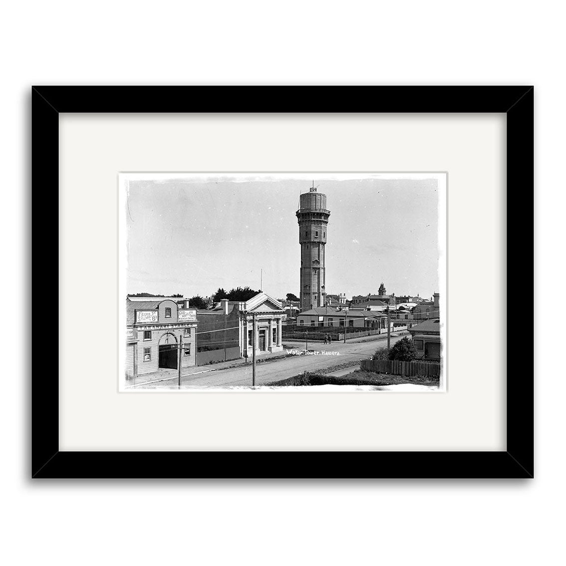 Archival black and white photo print Water Tower Hawera 1915 - HI150 shows a New Zealand city street with a tall cylindrical water tower, low buildings, and clear sky, framed in black with a white mat.