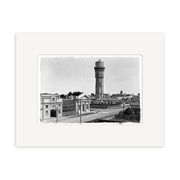 This framed archival print, Water Tower Hawera 1915 - HI150, features a black-and-white photo of a tall water tower with low buildings, scattered trees, and utility poles along a street, all displayed with a wide white mat.