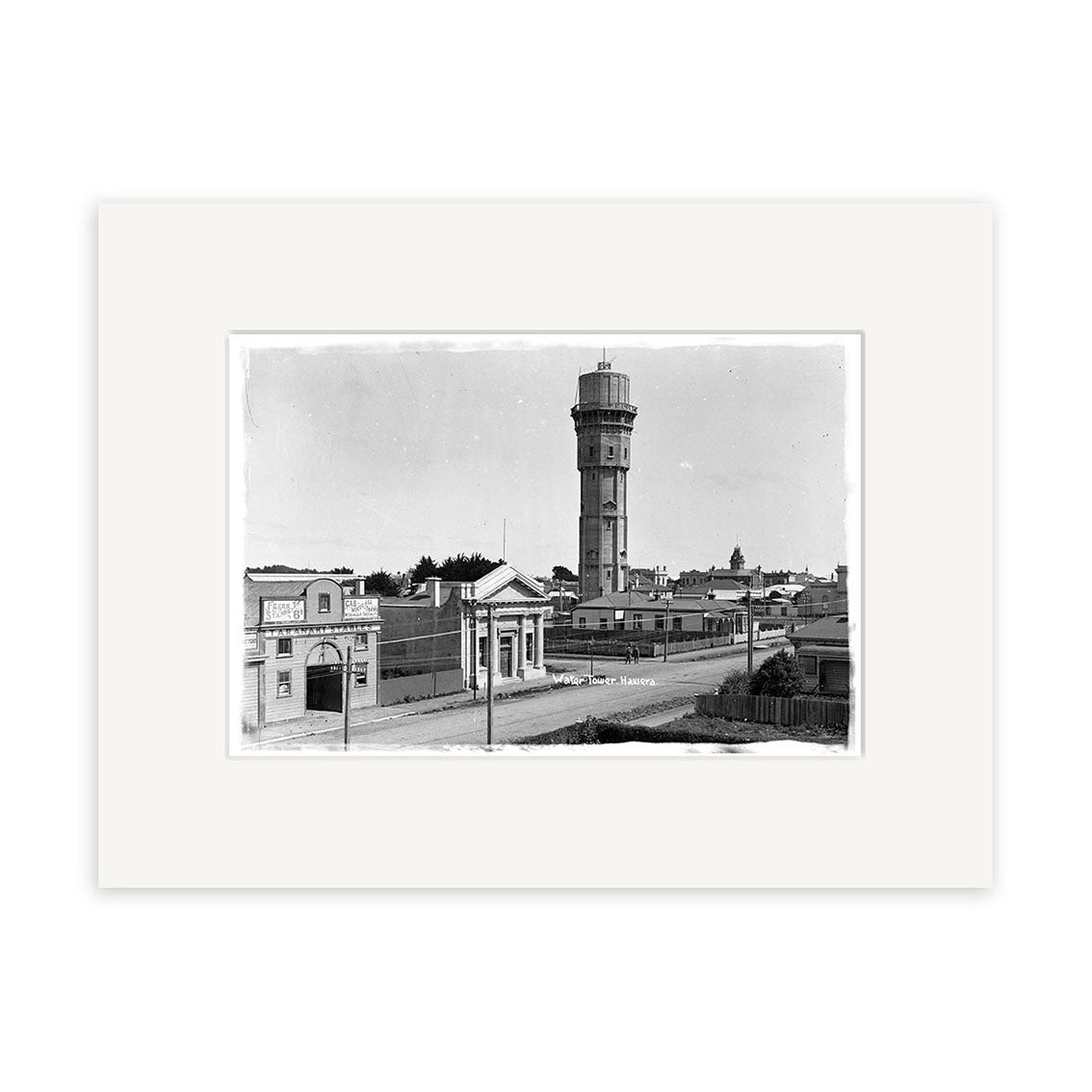 This framed archival print, Water Tower Hawera 1915 - HI150, features a black-and-white photo of a tall water tower with low buildings, scattered trees, and utility poles along a street, all displayed with a wide white mat.