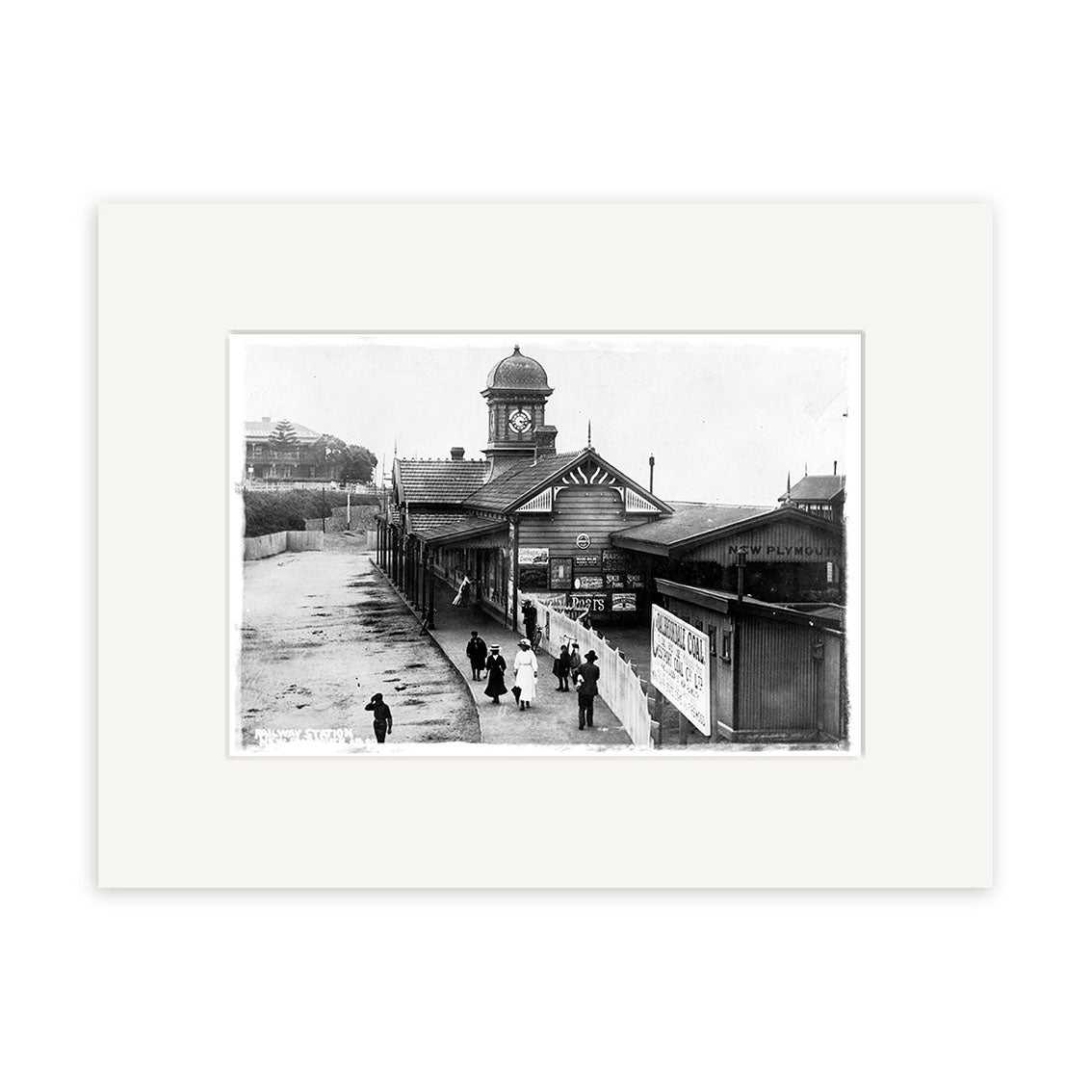 The New Plymouth Railway Station 1900 - HI151 is a black and white historical print showing a platform with people, a station building with clock tower and wall ads. Printed on archival matte paper, made in New Zealand.