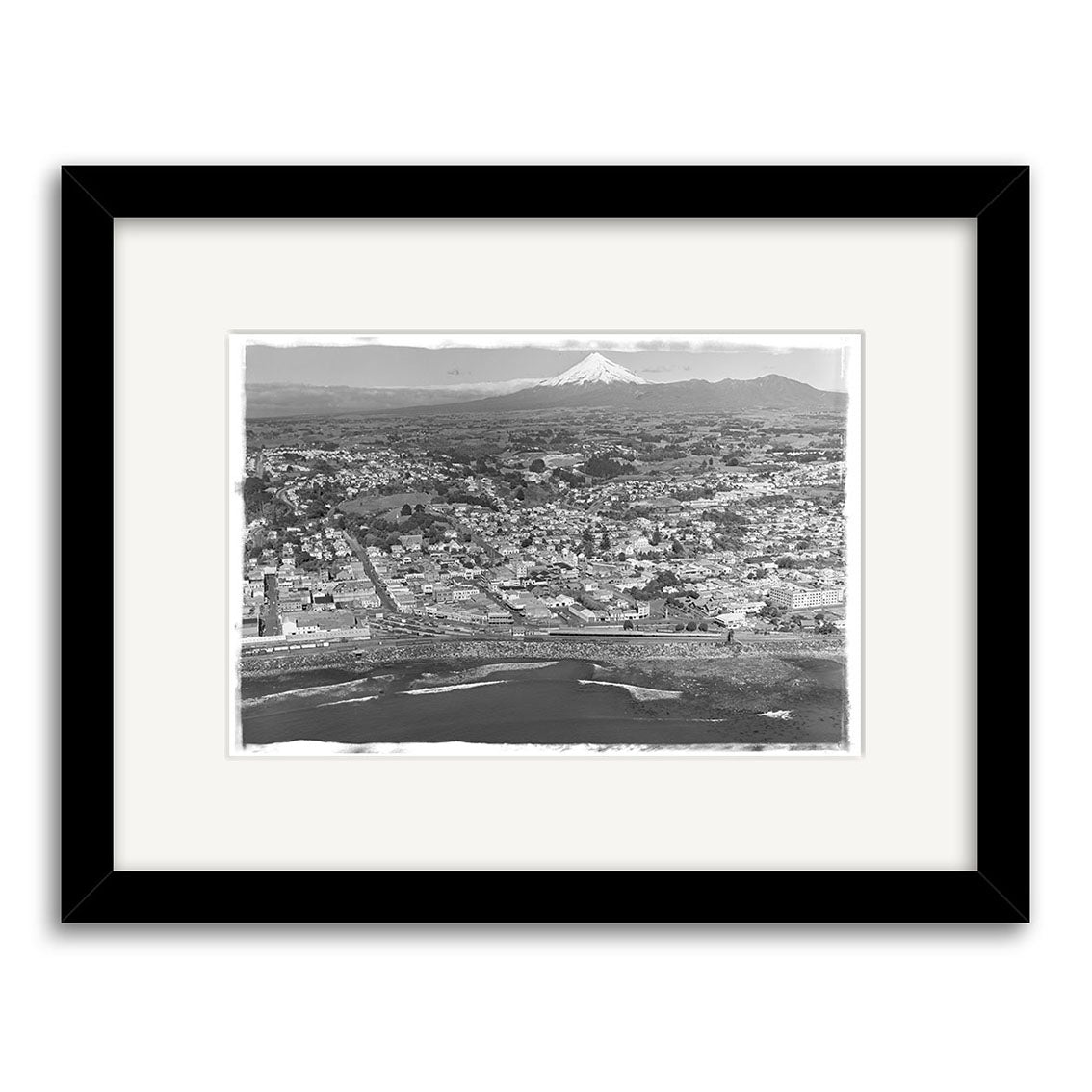 Taranaki Aerial 1951 - HI152: Black and white aerial photo of a coastal city, urban buildings, and a distant snow-capped mountain. This historical print is framed in black with a white mat on archival-quality matte paper.