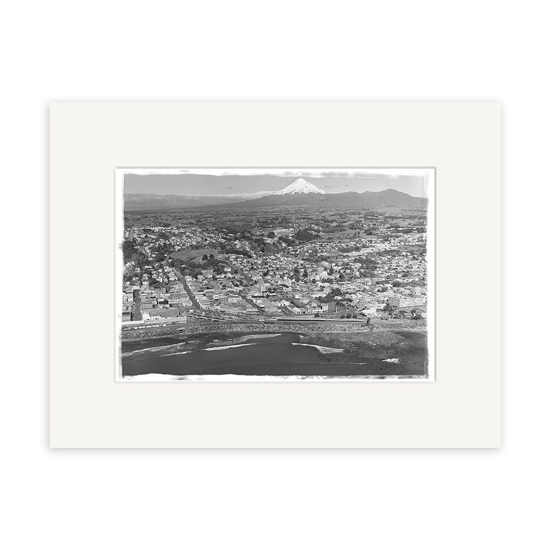 A black and white aerial matte print, Taranaki Aerial 1951 - HI152, shows a coastal city’s grid of streets and shoreline with a snow-capped mountain in the background in this historical photograph.