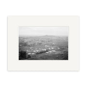 Black-and-white historical photographic print, Epsom 1910 – HI155, shows a New Zealand town with scattered houses and fields, hills in the background, and is framed with a white mat. The elevated view emphasizes its archival landscape quality.