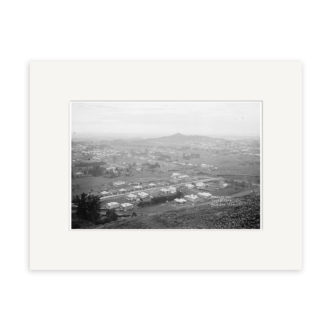Black-and-white historical photographic print, Epsom 1910 – HI155, shows a New Zealand town with scattered houses and fields, hills in the background, and is framed with a white mat. The elevated view emphasizes its archival landscape quality.