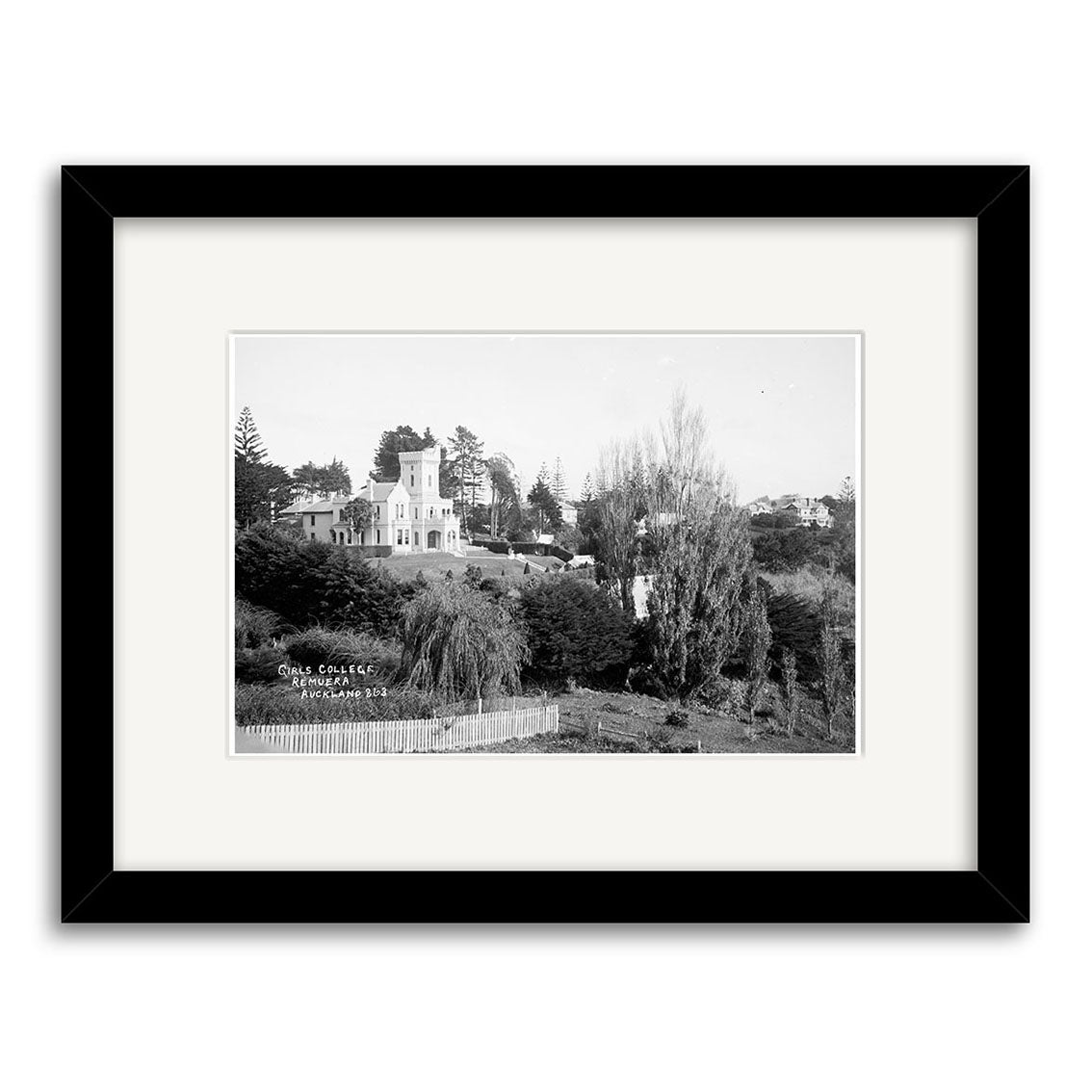A black-and-white historical photographic print, Girls College 1910 - HI156, depicts a large house with a tower in New Zealand, surrounded by trees and a wooden fence, framed with a wide white mat and black archival-quality frame.
