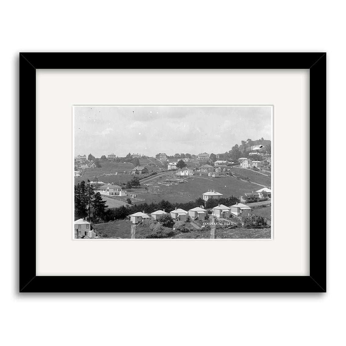 The Remuera 1910 No. 1 – HI157 is a black and white framed historical photographic print showing small houses, open fields, and trees in a hillside New Zealand neighborhood, capturing archival quality from the era.