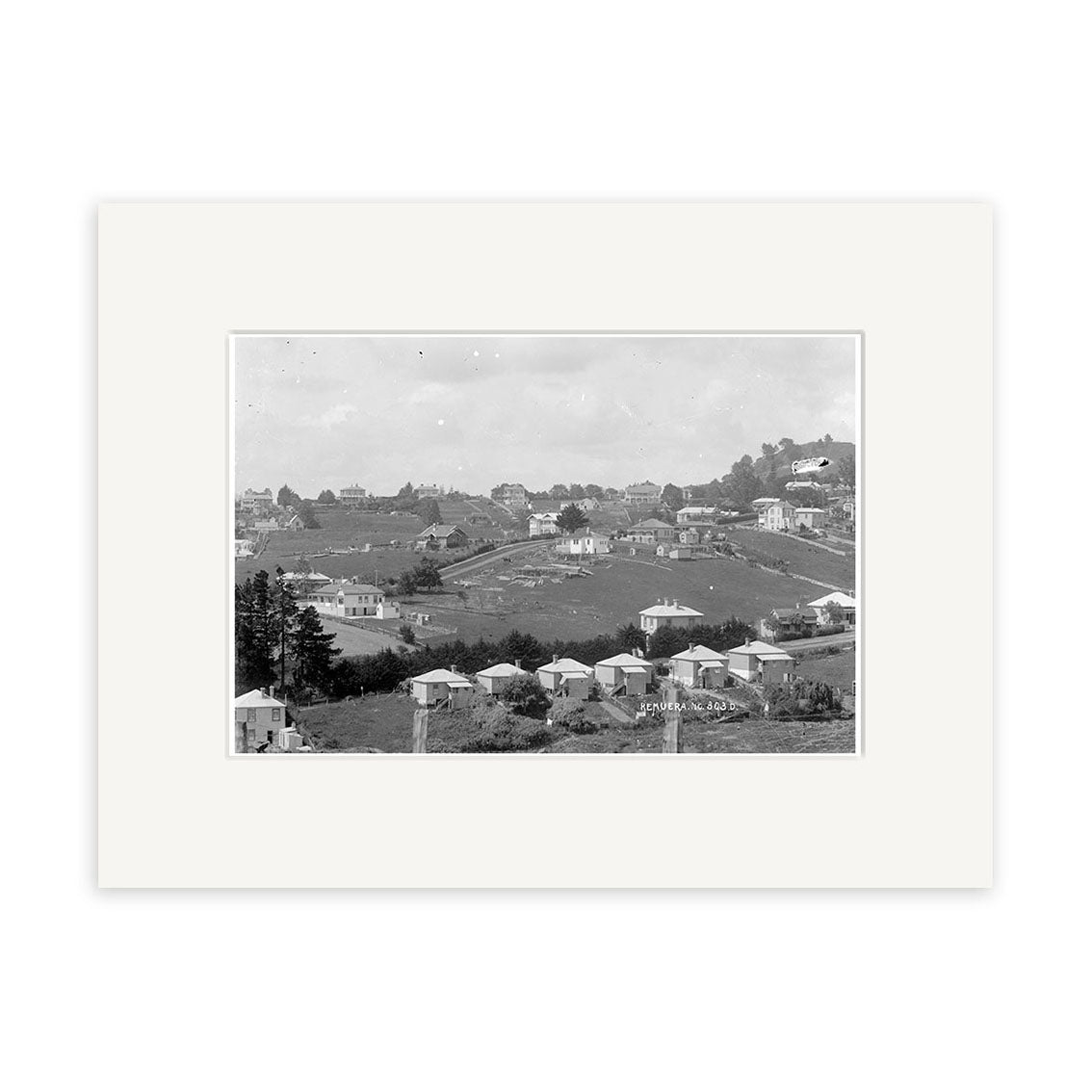 An archival quality black and white photograph, Remuera 1910 No. 1 - HI157, depicts a hilly residential area in New Zealand with scattered houses, trees, open land, and distant buildings beneath a cloudy sky.