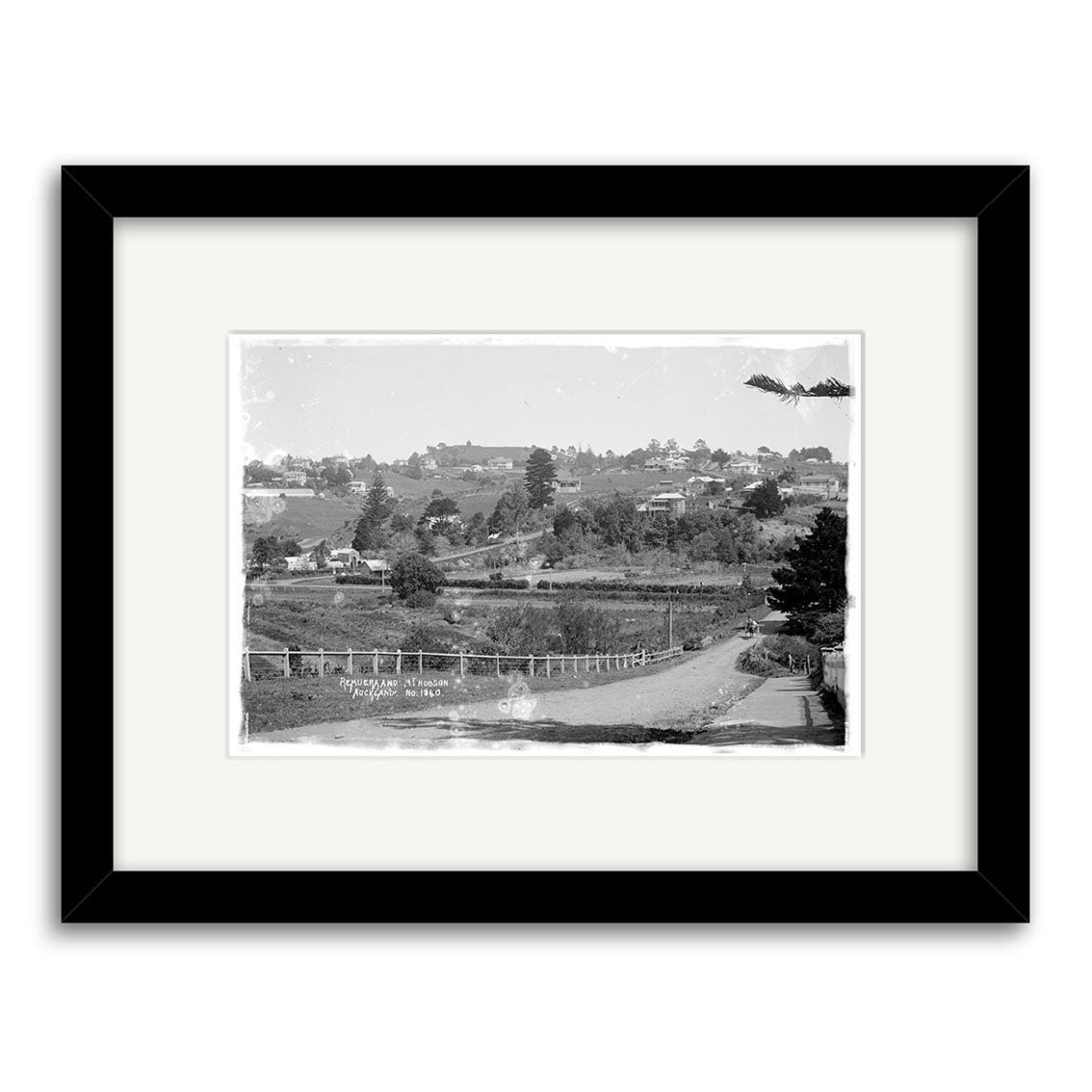 Framed black-and-white photographic print titled Remuera 1910 No. 2 - HI158 showing a rural New Zealand landscape with a dirt road, fence, trees, distant hillside buildings, and visible scratches and wear.