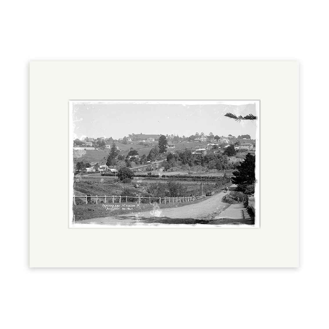 Black and white photograph titled Remuera 1910 No. 2 - HI158 featuring a rural New Zealand landscape with a dirt road, wooden fences, scattered trees, and houses under a cloudy sky. Archival quality print matted with a white border.
