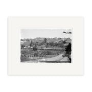 Black and white photograph titled Remuera 1910 No. 2 - HI158 featuring a rural New Zealand landscape with a dirt road, wooden fences, scattered trees, and houses under a cloudy sky. Archival quality print matted with a white border.