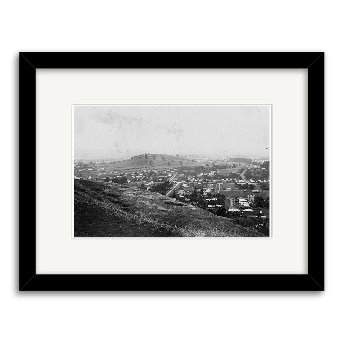 Epsom 1907 No.2 - HI159: Archival black-and-white photo print in a black frame of a hillside view overlooking a New Zealand town with scattered buildings, open fields, and a distant hill under a hazy sky.