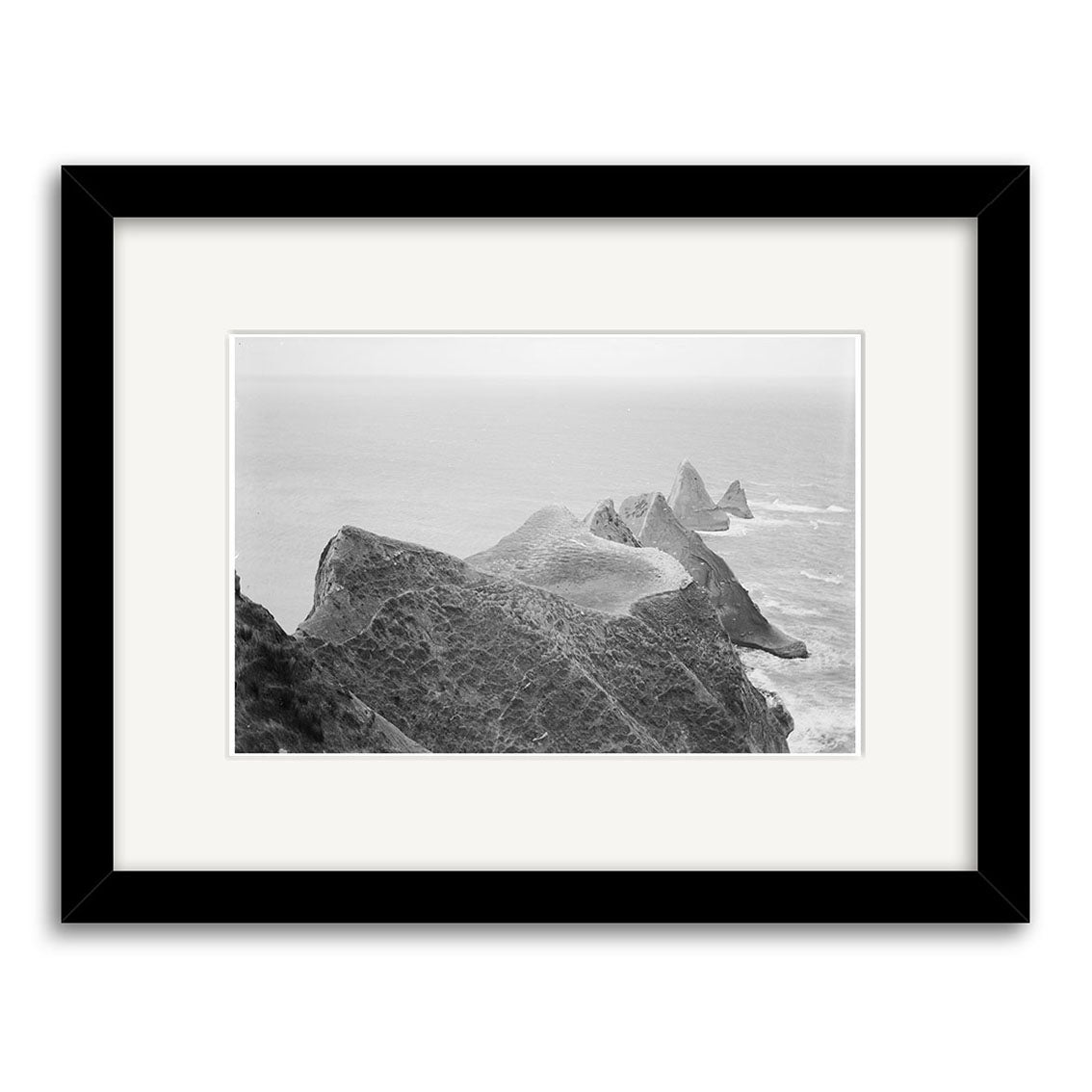 ‘Cape Kidnappers 1880 - HI161’: Black and white archival matte print of rocky cliffs extending into the ocean, misty headlands fading into the distance. Presented in a black frame with white mat.