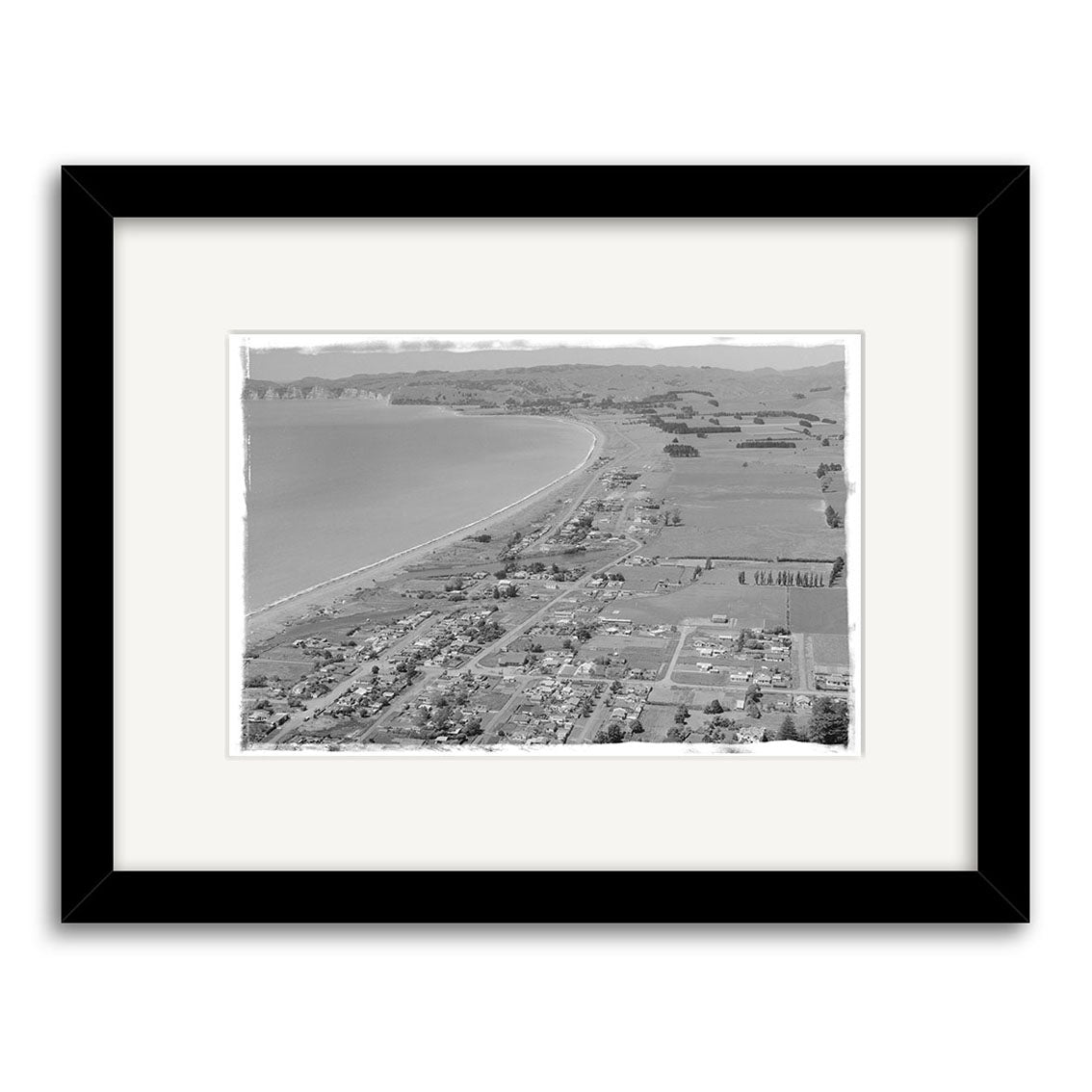 Black and white aerial photograph, Hawkes Bay 1958 - HI164. Shows a New Zealand coastal town with fields and homes by a long beach. Archival quality print with wide white mat and black frame.