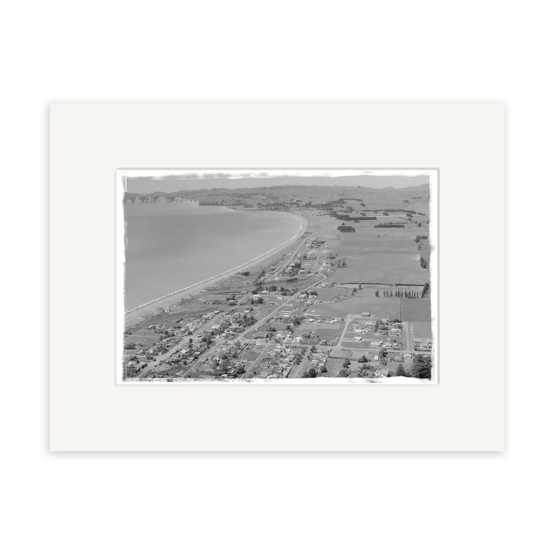 The “Hawkes Bay 1958 – HI164” is a black-and-white aerial photo of a New Zealand coastal town, showcasing a long beach, homes, fields, and distant hills. This historical print is mounted in an archival quality wide white mat border.