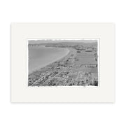 The “Hawkes Bay 1958 – HI164” is a black-and-white aerial photo of a New Zealand coastal town, showcasing a long beach, homes, fields, and distant hills. This historical print is mounted in an archival quality wide white mat border.