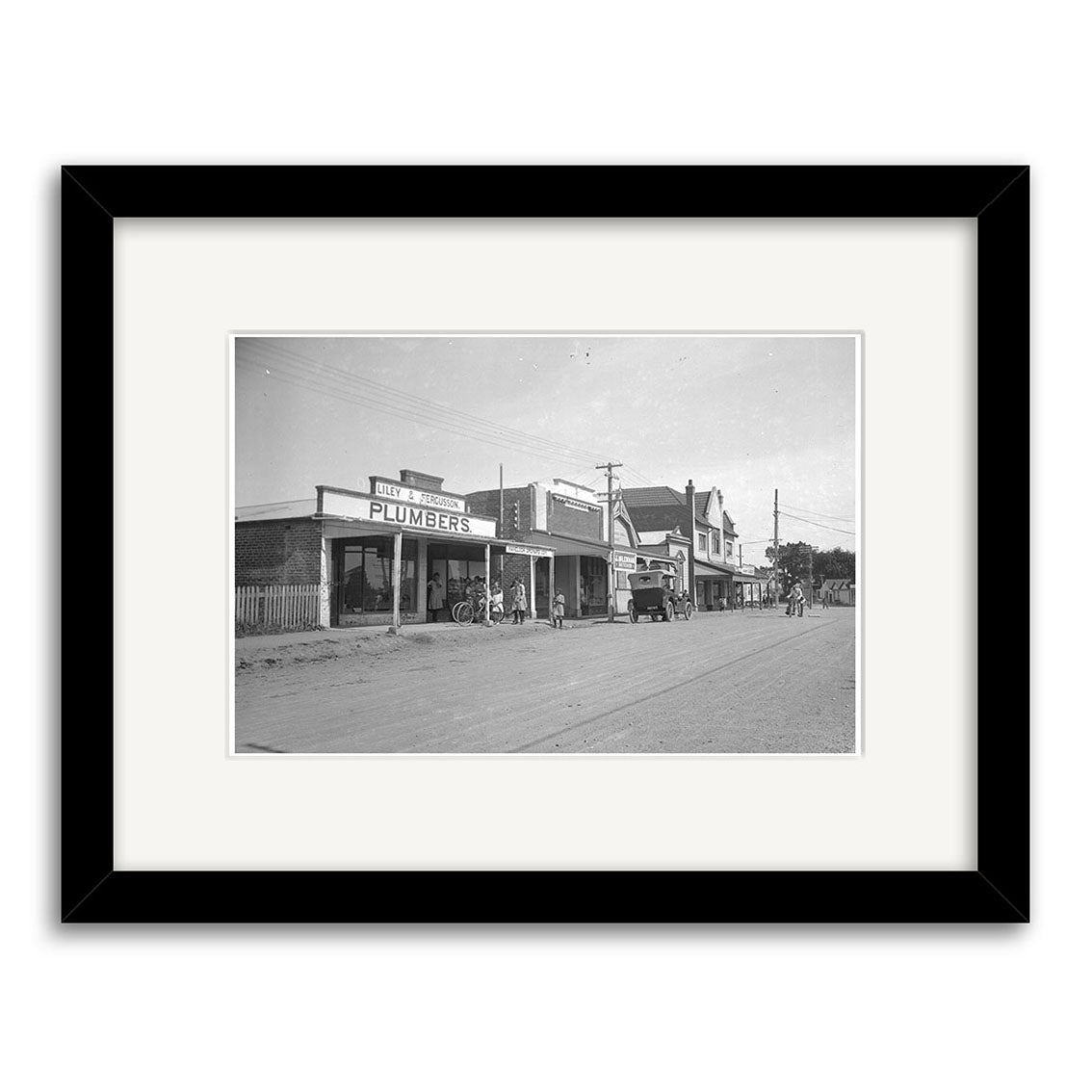 Black and white photo titled Havelock North 1925 - HI165 shows a small town street with a Plumbers sign, bicycles, people, and an early car on an unpaved road. Made in New Zealand on archival quality paper.