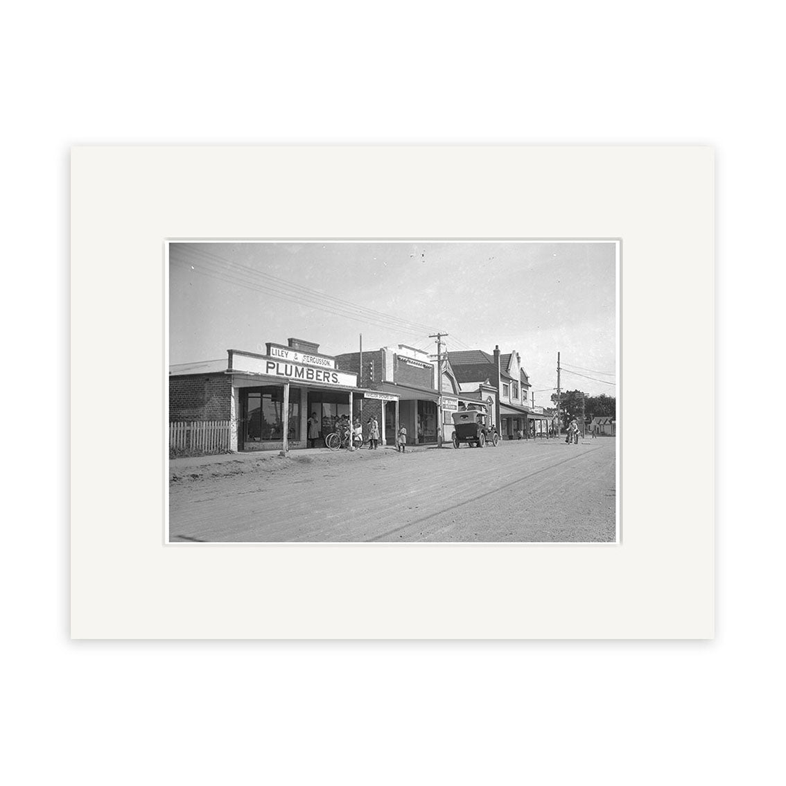 Black-and-white photo titled Havelock North 1925 - HI165 shows a small town street in New Zealand with storefronts, including a plumber’s shop, parked cars, bicycles, and people on the sidewalk; historical photographic print.