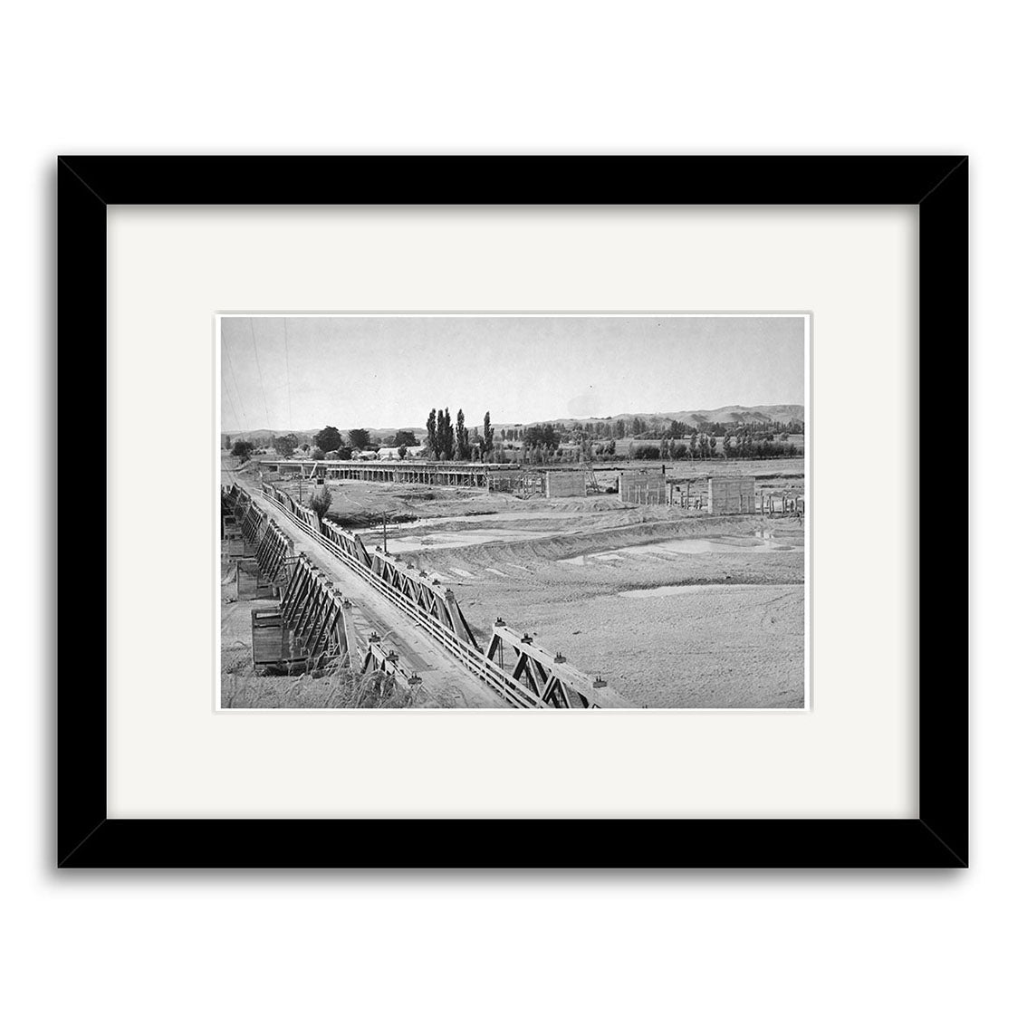Black and white photo titled Hawkes Bay 1948 - HI166 shows a long wooden aqueduct over dry land with people nearby, distant buildings, and trees. Printed on matte paper and archivally framed in black.