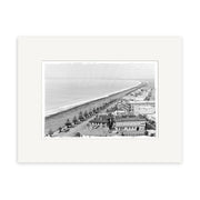 Archival quality black and white photo titled Napier 1890 - HI168, showing a coastal New Zealand city with a long beach, buildings, and a tree-lined road parallel to the shoreline, stretching into the distance.