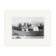 Black and white photo titled Bath Houses 1915 - HI169 shows two small gable-roofed buildings amid grass, trees, and mountains. This historical New Zealand print is mounted with a wide white mat.