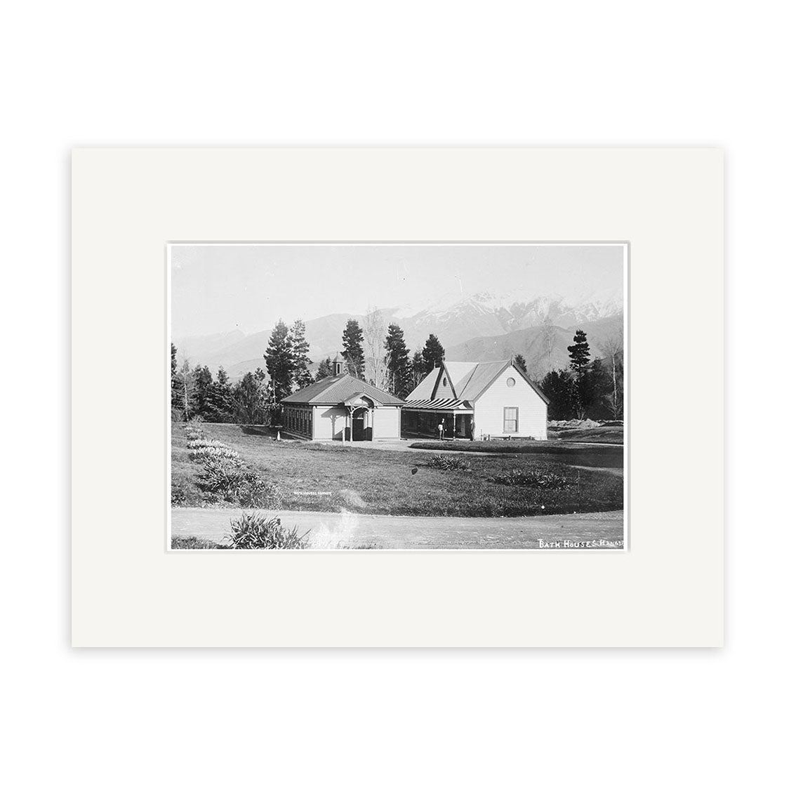 Black and white photo titled Bath Houses 1915 - HI169 shows two small gable-roofed buildings amid grass, trees, and mountains. This historical New Zealand print is mounted with a wide white mat.