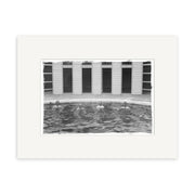 The black and white print Baths Hanmer Springs 1900 - HI170 depicts four people partially submerged in a circular outdoor pool before a building with tall windows or doors, bordered by a white frame.