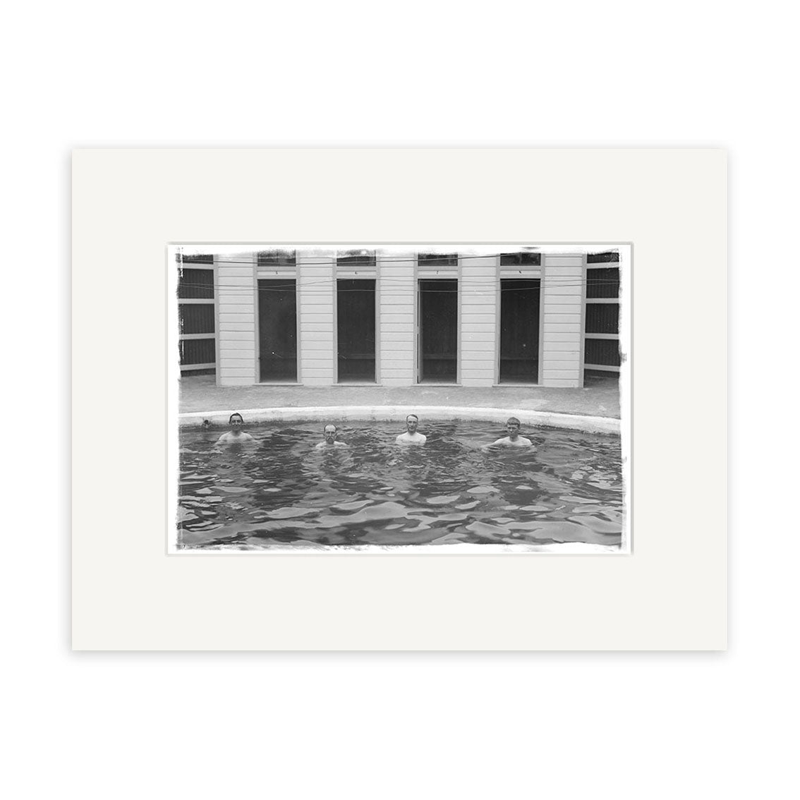 The black and white print Baths Hanmer Springs 1900 - HI170 depicts four people partially submerged in a circular outdoor pool before a building with tall windows or doors, bordered by a white frame.