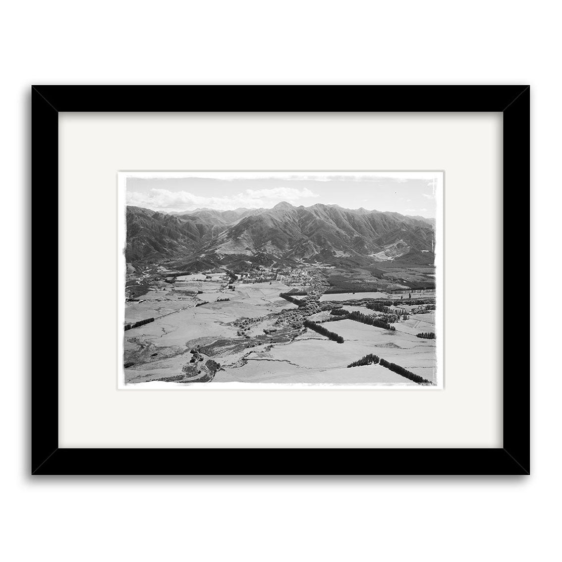 A black-and-white aerial photo titled Hanmer 1959 - HI172 shows open fields, a small town, and mountains. Presented as a Historical Photographic Print on archival matte paper in a black frame with white mat.