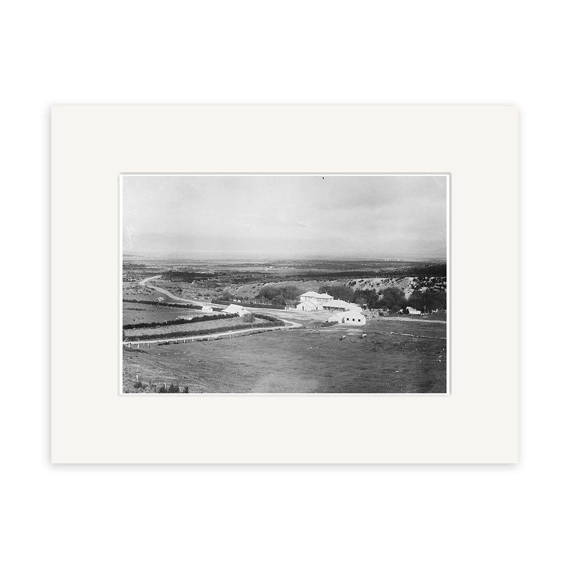 Black and white photo titled Jollies Pass hotel 1915 - HI173 shows a rural New Zealand landscape with a winding road, fenced fields, and distant hills. This archival quality print is presented with a white mat.
