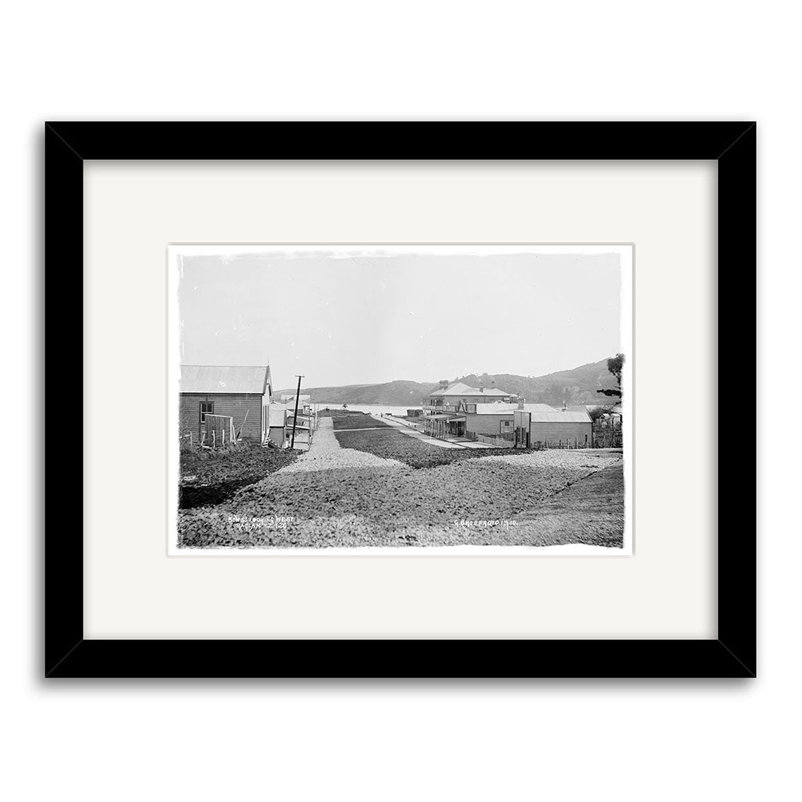 Black-and-white photo titled Bow Street Raglan 1910 - HI174, showing a dirt road lined with buildings and hills beyond. This historic print comes beautifully framed with a white mat and a sleek black frame.