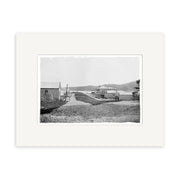 The Bow Street Raglan 1910 - HI174 print features a black-and-white photo on archival paper of a dirt road with wooden buildings and hills under a clear sky, centered on a white mat for a timeless historical display.