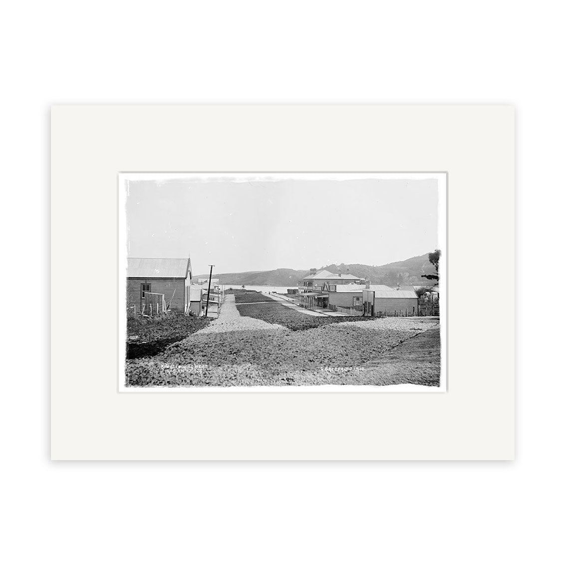 The Bow Street Raglan 1910 - HI174 print features a black-and-white photo on archival paper of a dirt road with wooden buildings and hills under a clear sky, centered on a white mat for a timeless historical display.