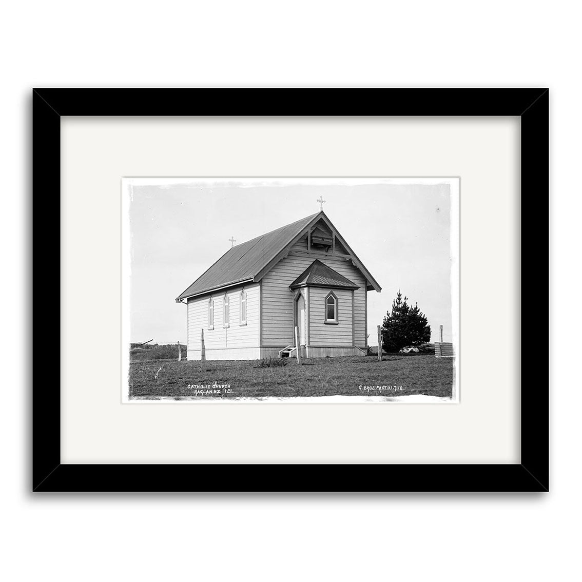 This archival-quality black-and-white print, Catholic Church 1910 - HI175, shows a small wooden church with a steep metal roof and cross, set on grass with a tree to the right. Framed in black with a white mat. Made in New Zealand.