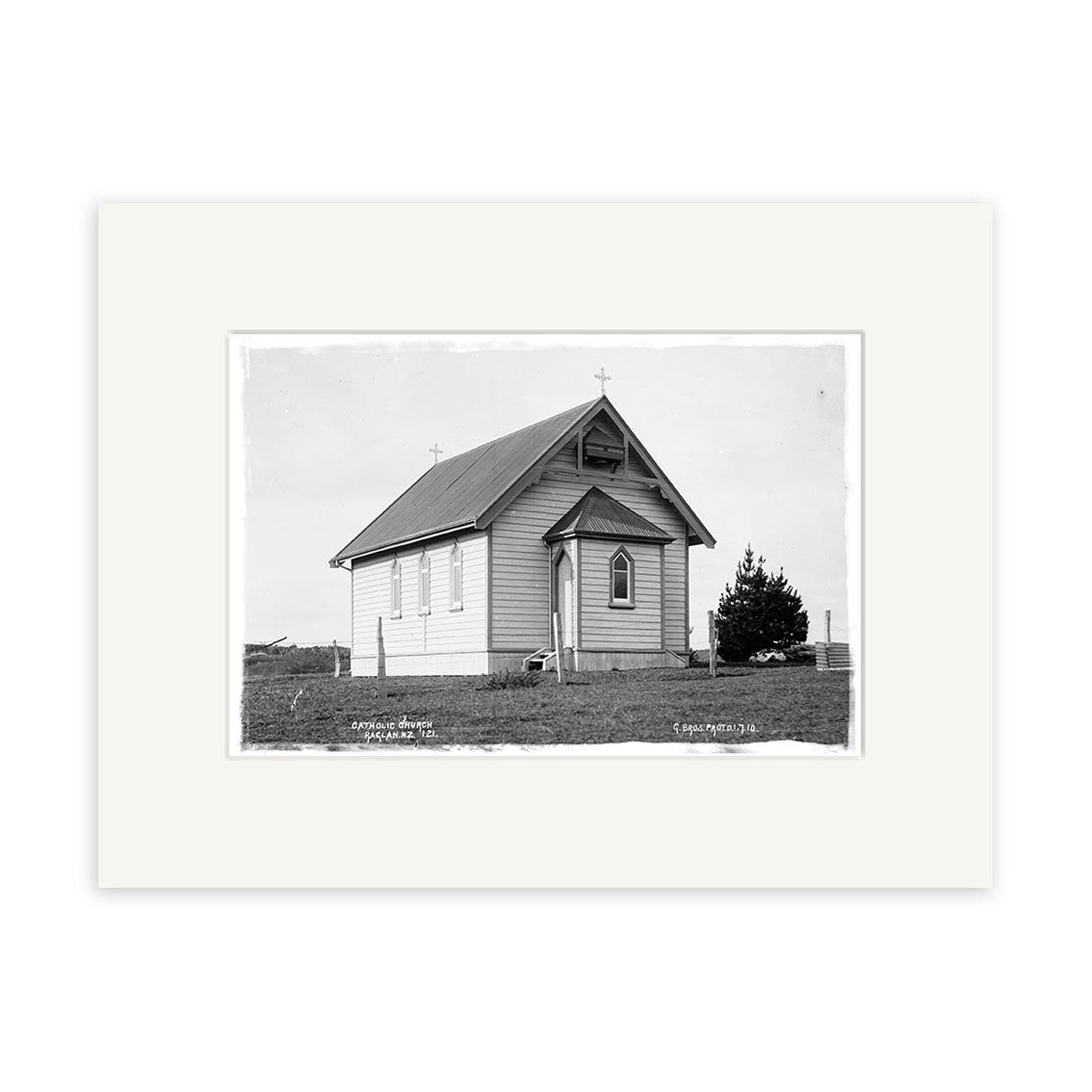 Archival-quality black and white photographic print of Catholic Church 1910 - HI175: a small wooden church with a steep metal roof, front porch, and crosses on top, standing on a grassy lot with a single tree. Made in New Zealand.