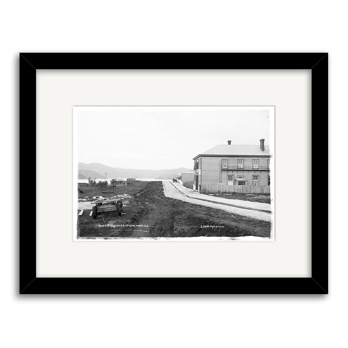 The Cliff Street 1910 - HI176 print shows a dirt road, old car, and two-story building in New Zealand with hills beyond. Presented as a black and white historical photo, it comes framed with a white mat and black frame.