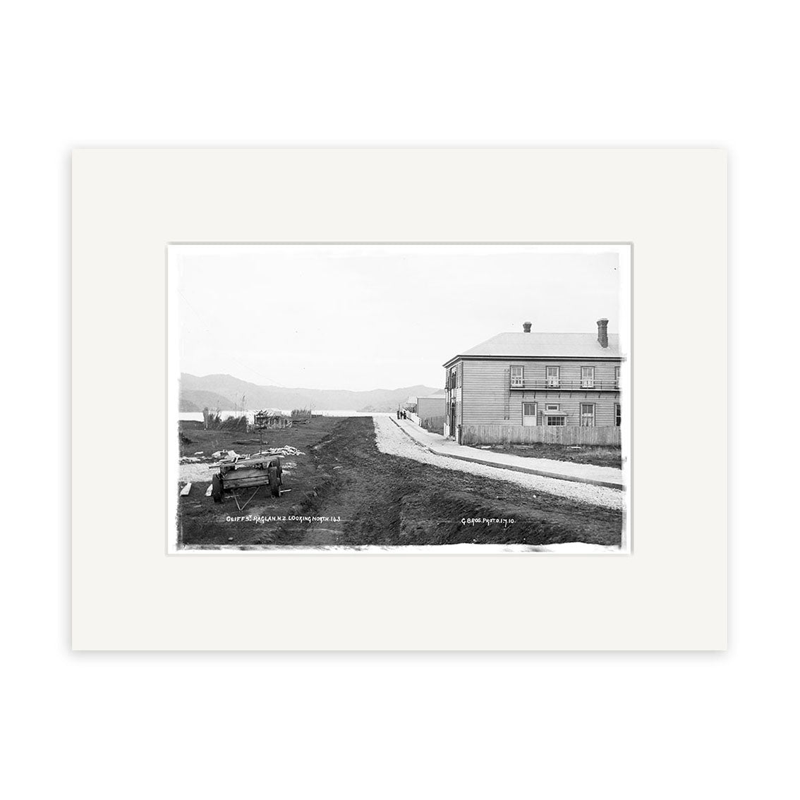 Cliff Street 1910 - HI176: Black and white historical print of a dirt road with a horse-drawn carriage and large two-story building in rural New Zealand hills, on archival quality matte paper for lasting detail.