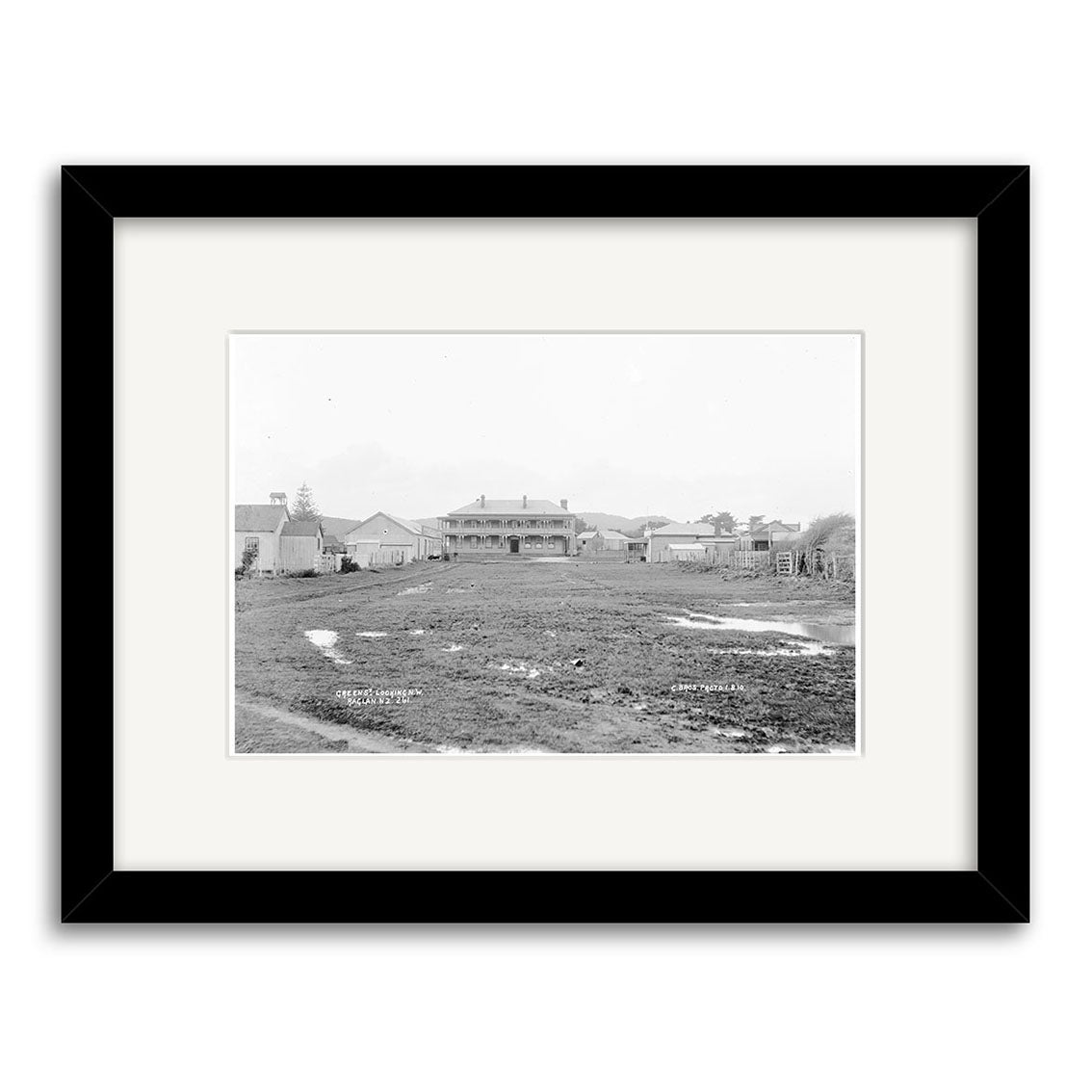 Black-and-white photo Green St Rangiora 1910 - HI178 shows a large New Zealand building with veranda, smaller structures, and muddy field under overcast sky. Historical print presented in black frame with archival-grade matte paper.