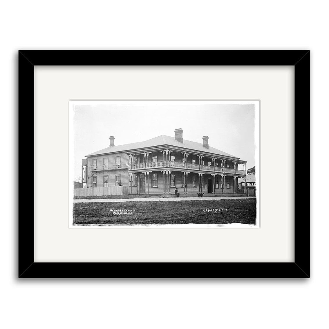 Archival-quality black-and-white photographic print of the Harbour View Hotel 1910 (HI179), a two-story wooden building in New Zealand with wraparound veranda, decorative posts, chimneys, and people on the porch and lawn; framed with white mat.