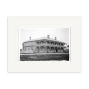 Archival quality black and white photo print of Harbour View Hotel 1910 (HI179), showing a two-story wooden building with wraparound porch, chimneys, people on the steps, and a picket fence in New Zealand. Mounted with white mat.